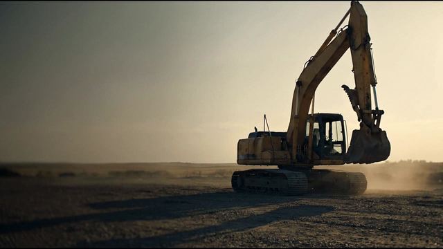 A tracked excavator on open gravel with its boom and bucket raised and dust drifting behind it
